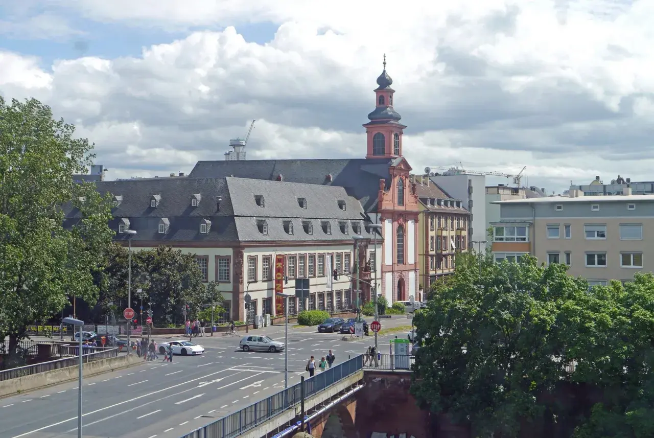 Barocke Kirche mit Turm und Mansarddach, Teil des Jakobsweg Hessen. Autos fahren auf einer Straße, Menschen überqueren sie.