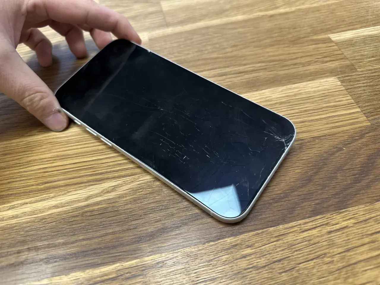 Dłoń trzyma telefon z pękniętym szkłem hartowanym. Ekran pokryty jest siecią drobnych pęknięć, szczególnie widocznych w prawym dolnym rogu.