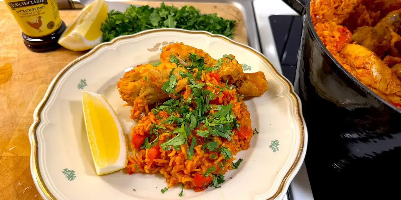 En tallrik med arroz con pollo, kyckling och ris med tomat, garnerad med persilja och citronklyfta. En gryta med samma rätt syns i bakgrunden.