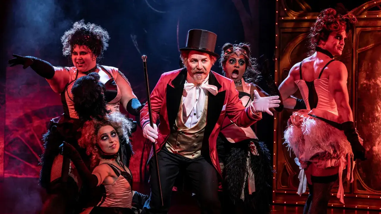 Scen från Moulin Rouge! The Musical i Stockholm. En cirkusdirektör med cylinderhatt och röd kavaj leder en färgstark ensemble.
