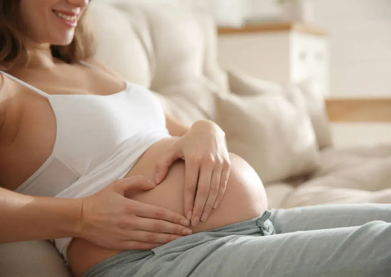 A pregnant woman rests on a couch, her hands forming a heart on her belly. She smiles, perhaps thinking about the baby, boy or girl, and the early pregnancy insomnia.