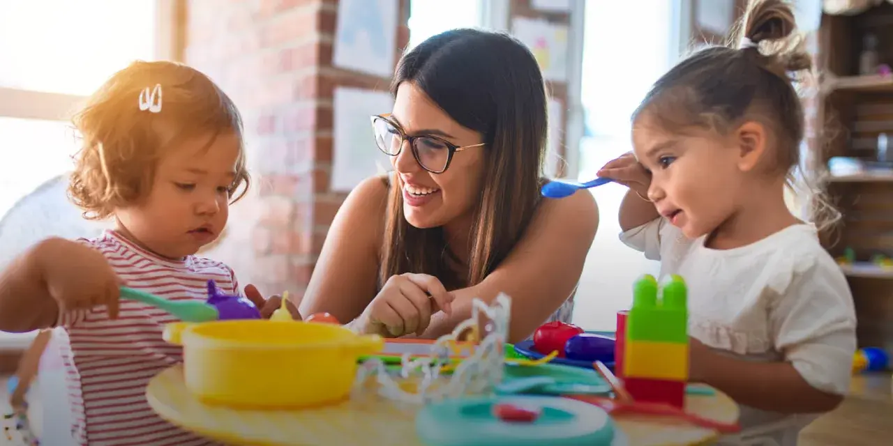 Eine lächelnde Frau mit Brille spielt mit zwei Kleinkindern. Sie lernt den erzieher berufsbegleitend und fördert spielerisch die Entwicklung der Kinder.