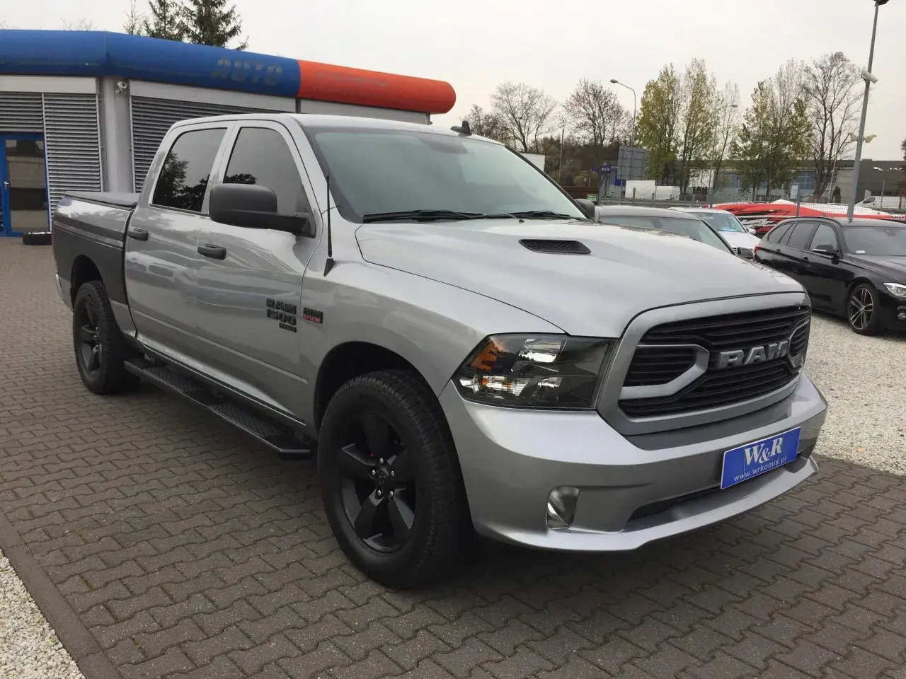 Srebrny Dodge Ram 1500 5.7 Hemi z czarnymi felgami i przyciemnianymi szybami stoi na parkingu przed budynkiem z niebiesko-pomarańczowym banerem.