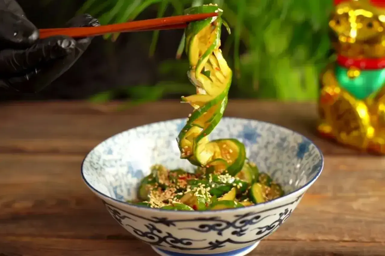 Chopsticks podnoszą zwinięty plasterek ogórka z miski pełnej tajskiej sałatki z ogórka, posypanej sezamem.