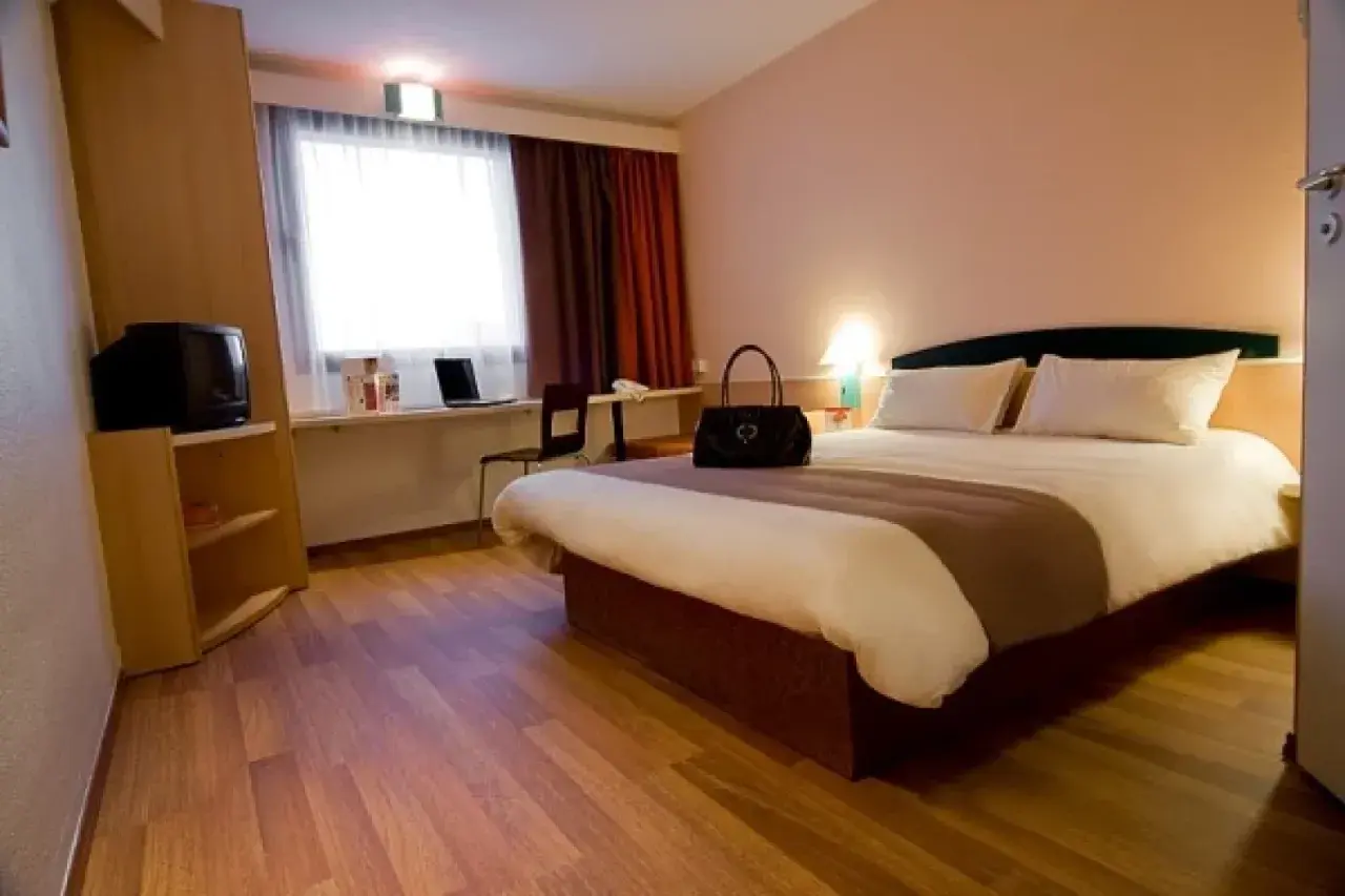 Pokój w Ibis Hotel Zabrze: łóżko, toaletka z laptopem, szafa z telewizorem i okno z zasłonami.