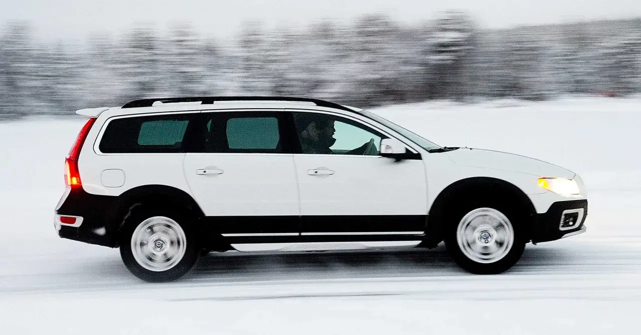 Vit Volvo XC70 kör på en snöig väg, med en förare bakom ratten. Bilen är i rörelse, vilket skapar en suddig bakgrund av träd.