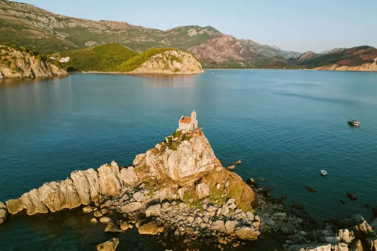A tiny church perches on a rocky islet, a picturesque stop on a Montenegro itinerary. Mountains rise behind the tranquil blue bay.
