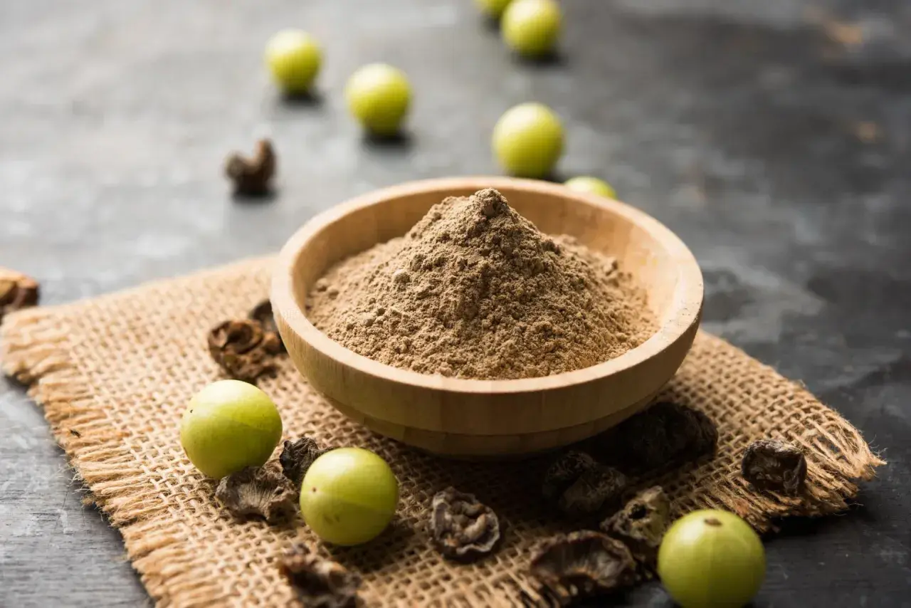 Polvere di triphala in una ciotola di legno, con accanto frutti di amla e parti essiccate. Il triphala a cosa serve? Aiuta la digestione e il benessere generale.