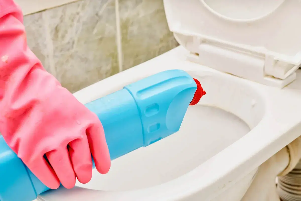 Mano con guanto rosa usa detergente per pulire il water incrostato.