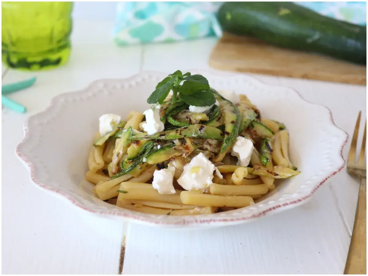 Pasta fredda con zucchine grigliate e cubetti di formaggio fresco, guarnita con erbe aromatiche. Un piatto estivo leggero e gustoso.