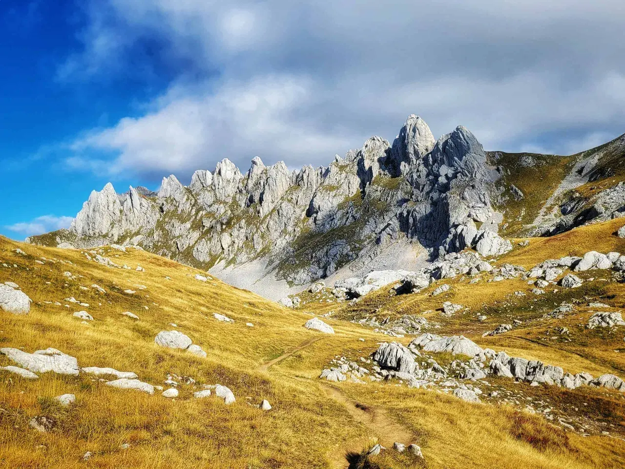 A winding path leads through golden grass and rocks towards the jagged peaks of Bobotov Kuk, a stunning hike under a dramatic sky.