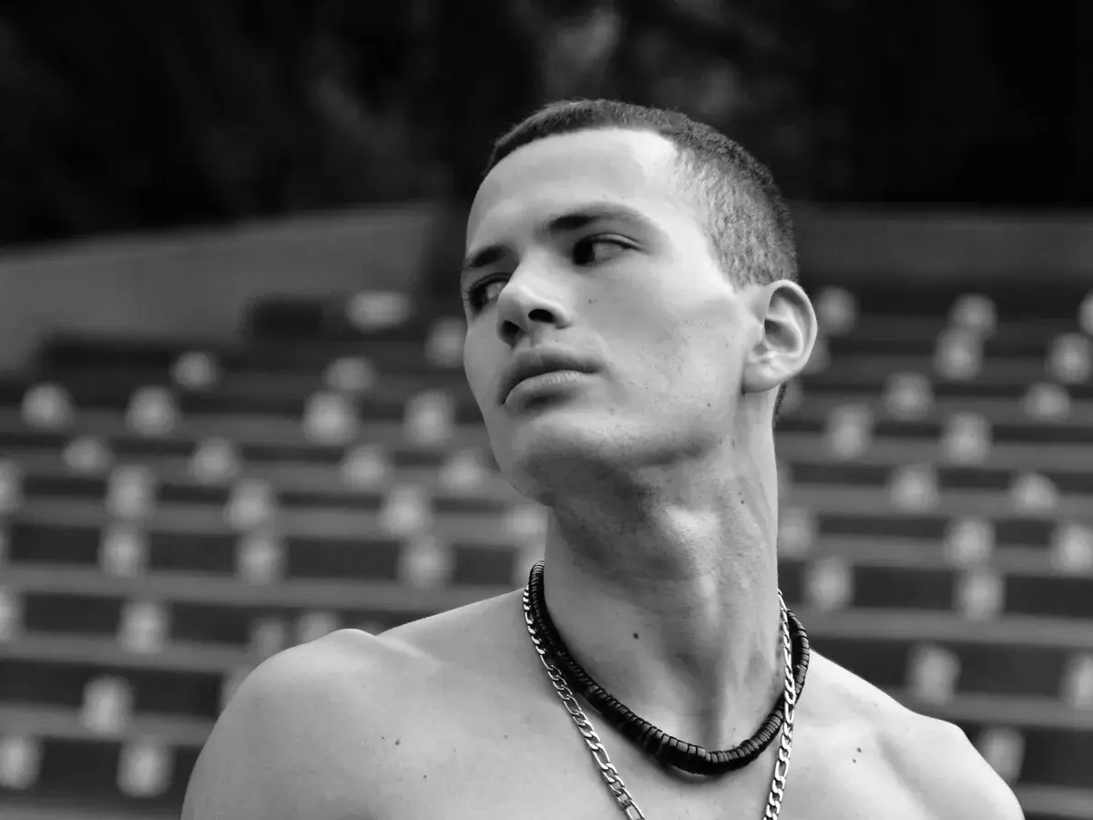 Joven con buzz cut largo, collar plateado y mirada perdida al horizonte.
