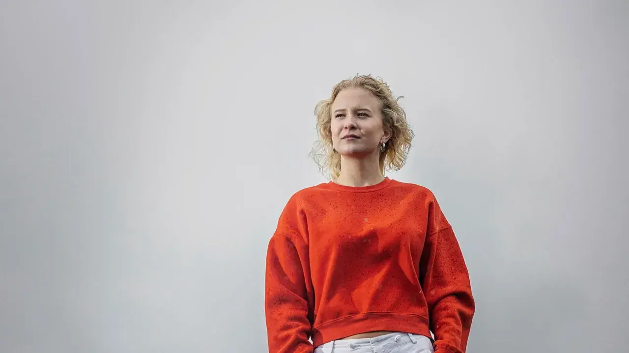 Sigrid Johnson, med sitt blonda, lockiga hår, bär en orange tröja och tittar drömmande bort.