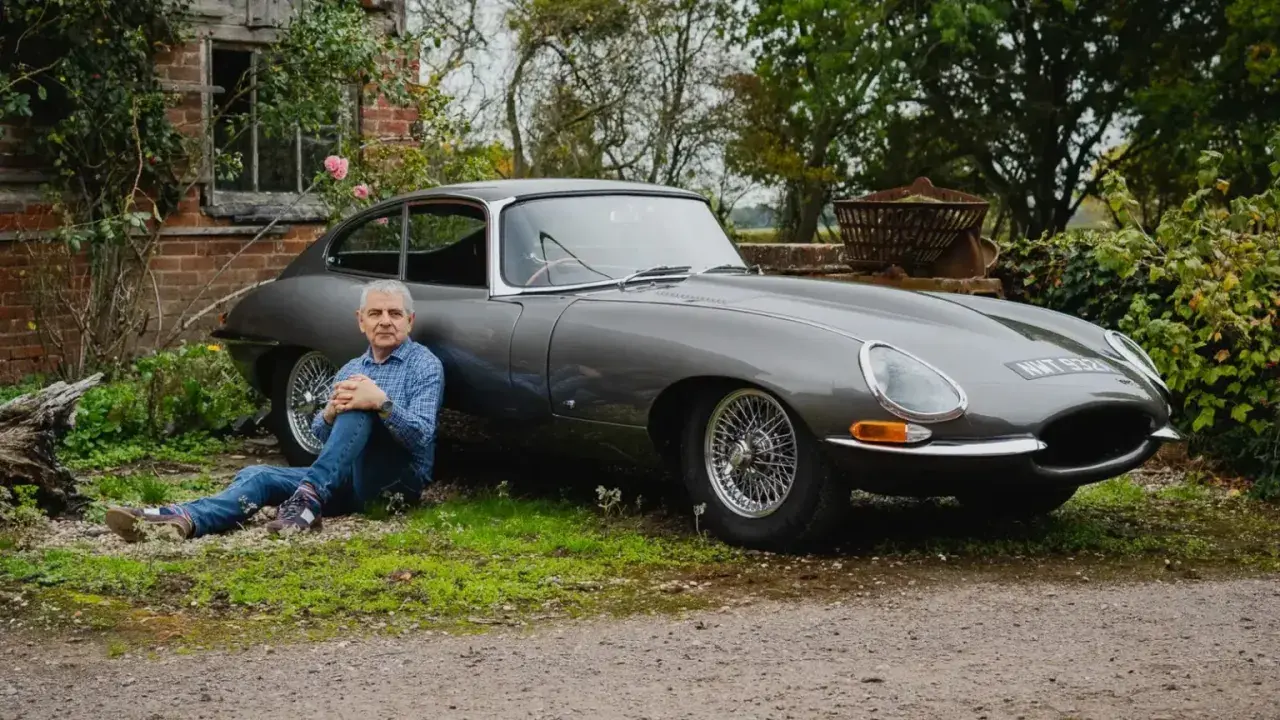 Man sitter vid en grå Jaguar E-Type i lantlig miljö.