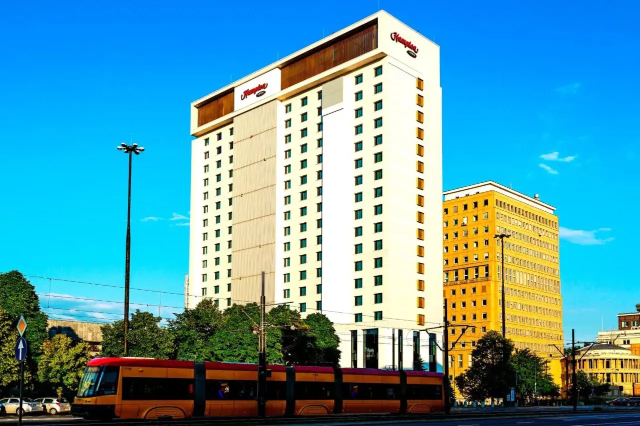 Hampton by Hilton Warsaw, nowoczesny hotel z charakterystycznym logo, góruje nad miejskim krajobrazem. Obok niego żółty budynek i tramwaj w ruchu.