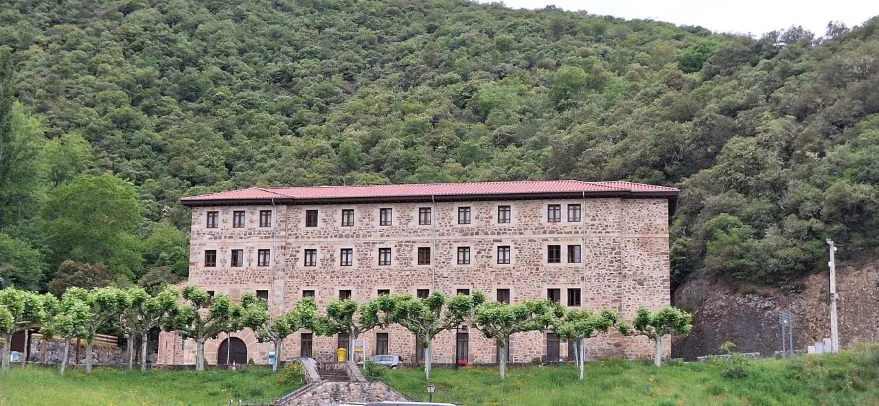 Edificio de piedra con tejado rojo, rodeado de árboles y un monte frondoso. Es el monasterio de Santo Toribio de Liébana.