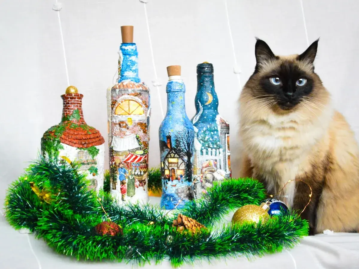 Urocze butelki ozdobione techniką decoupage na szkle, z zimowymi scenkami. Obok siedzi piękny kot.