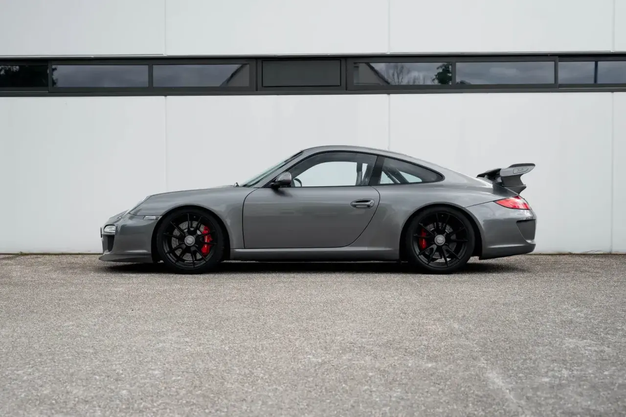 Grauer Porsche 997 GT3 RS mit schwarzem Flügel und roten Bremssätteln steht vor einer weißen Wand.