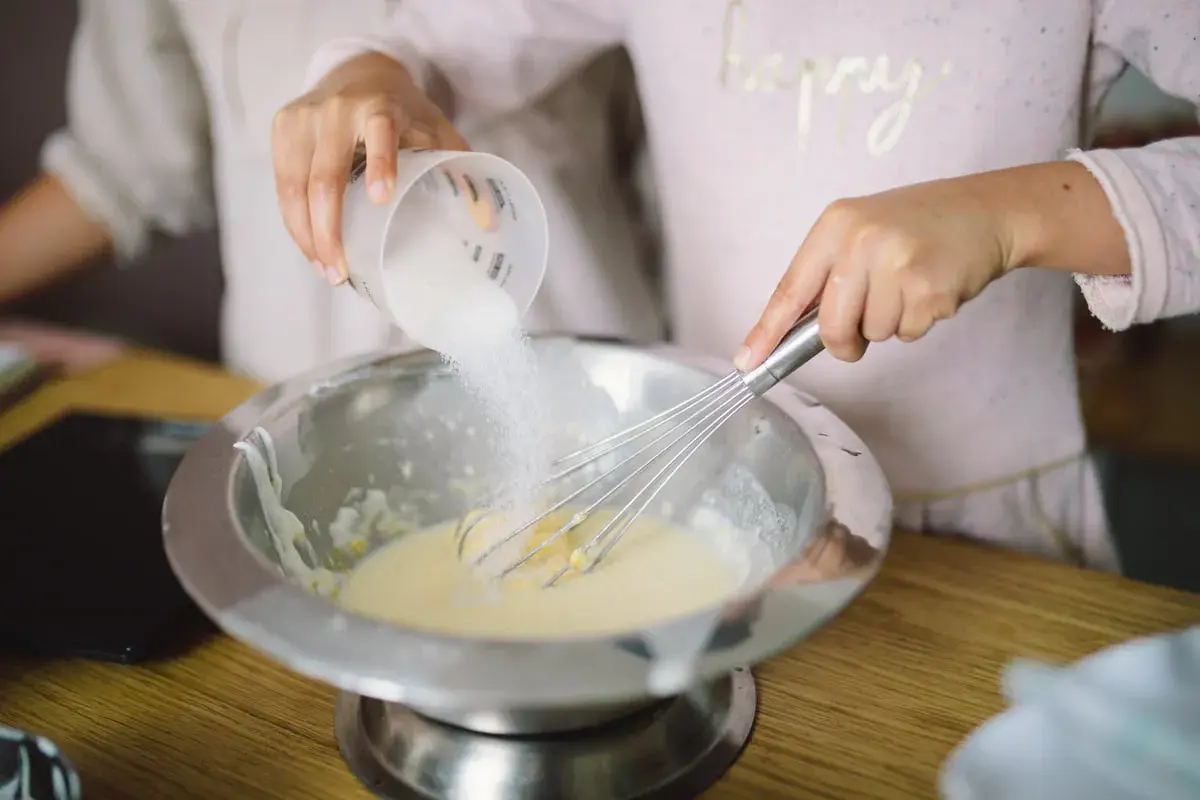 Préparation de la pâte à crêpes : du sucre est versé dans un saladier où une main fouette la préparation.