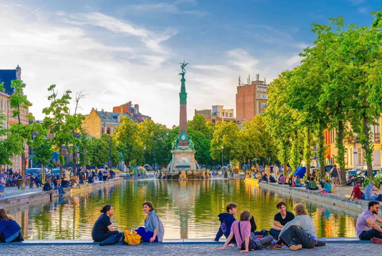 Des gens profitent d'une journée ensoleillée au bord d'un étang, un monument imposant se dresse en arrière-plan. Une belle scène pour visiter Bruxelles.