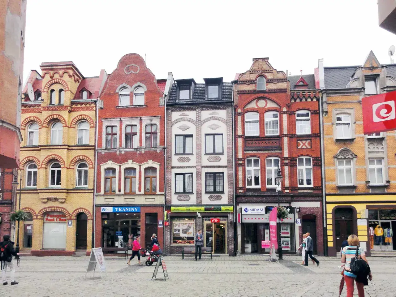 Kolorowe kamienice na lęborkim starym mieście, z witrynami sklepów i przechodniami na brukowanym placu.
