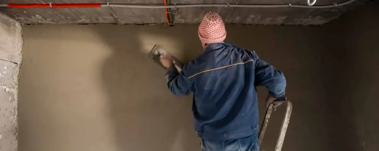 Mann mit Mütze verputzt eine Wand im Keller. Der **Sanierputz** im **Keller** wird sorgfältig aufgetragen.