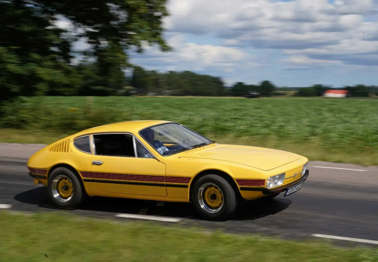 Gul vw sp2 susar fram längs en landsväg, med gröna fält och moln som suddas ut i bakgrunden.
