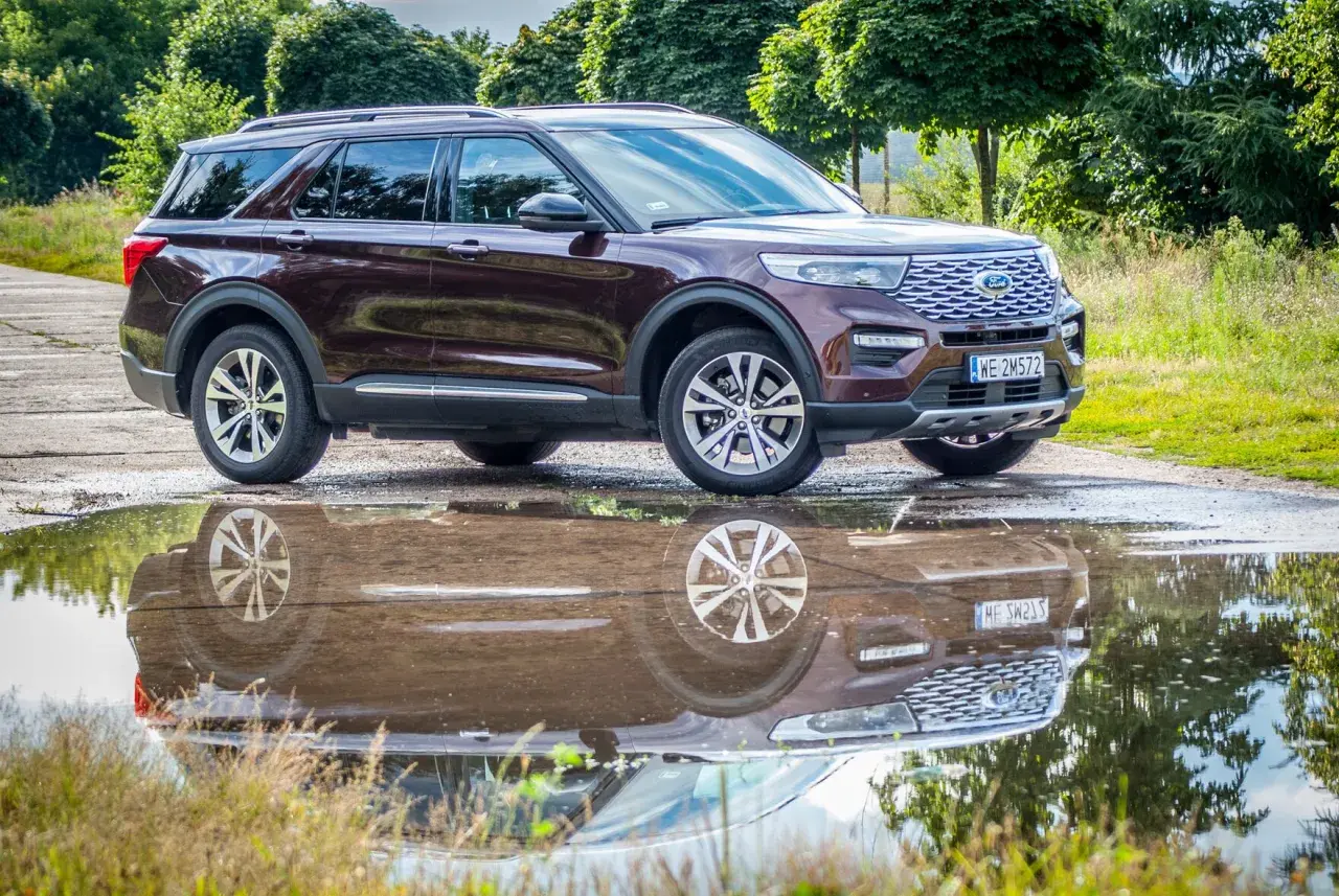 Ciemnobordowy Ford Explorer, jeden z popularnych ford suv modele, odbija się w kałuży po deszczu, otoczony zielenią.