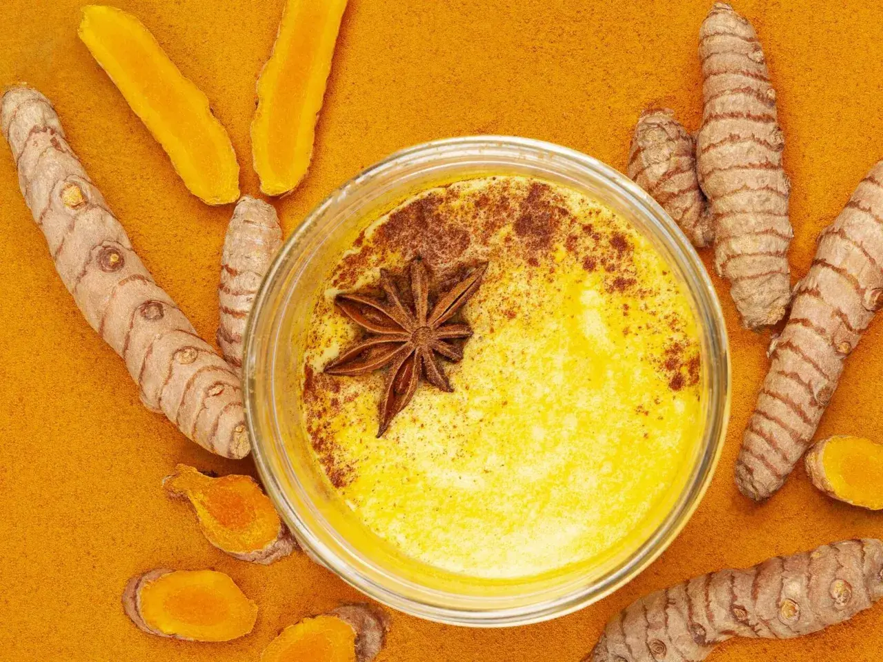 Goldene Milch mit Kurkuma und Honig, verziert mit Sternanis. Umgeben von frischen Kurkumawurzeln und Pulver.