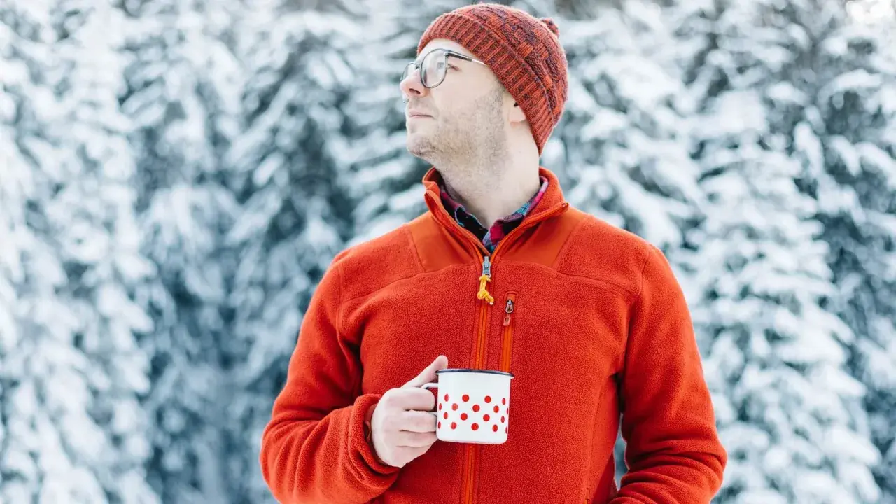 Mężczyzna w pomarańczowym polarze i czapce trzyma kubek w śnieżnym lesie. Polar jest wykonany z miękkiego, ciepłego materiału.
