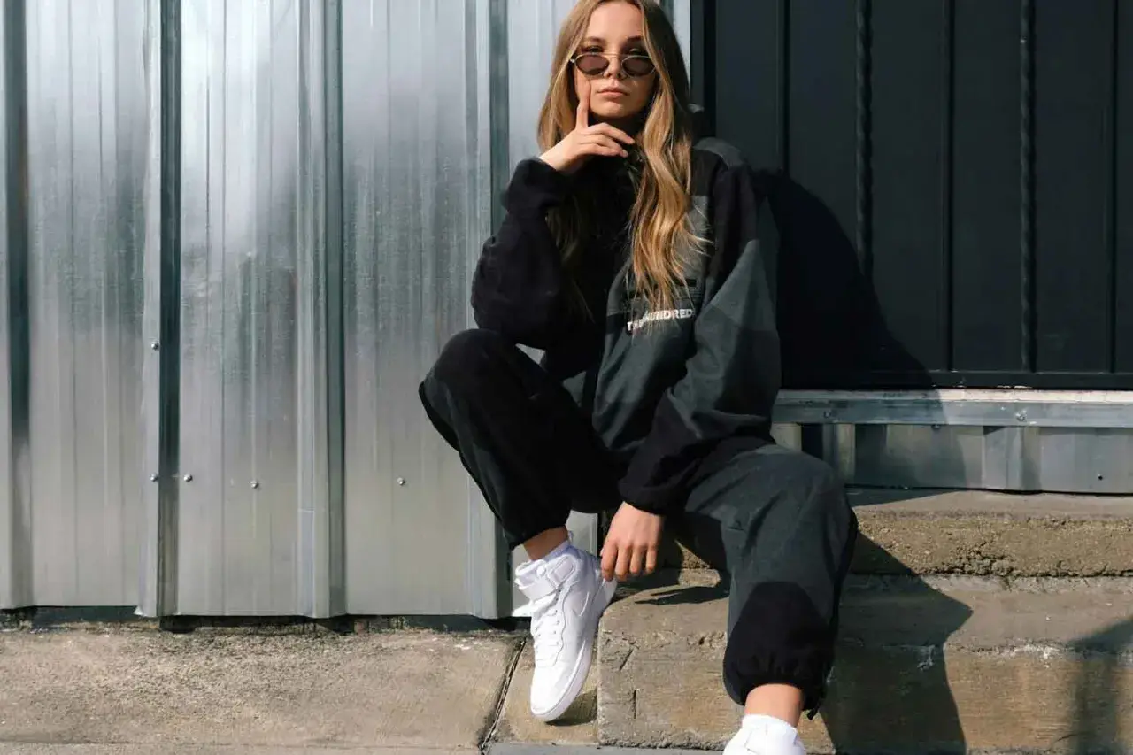 Chica con estilos de ropa mujer modernos, posa con sudadera y pantalones deportivos, zapatillas blancas y gafas de sol.