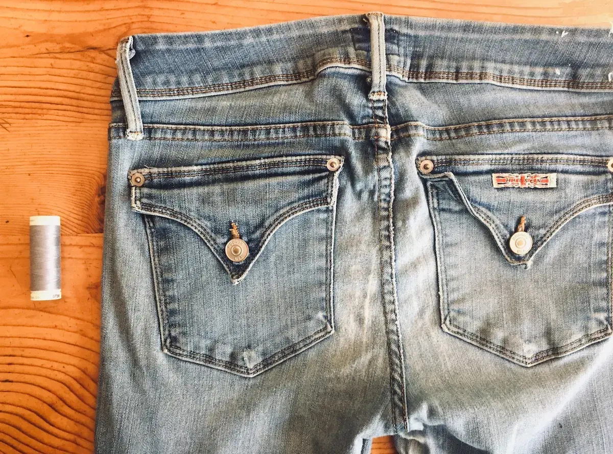 A spool of grey thread sits beside the back pockets of light blue jeans, ready for invisible mending.