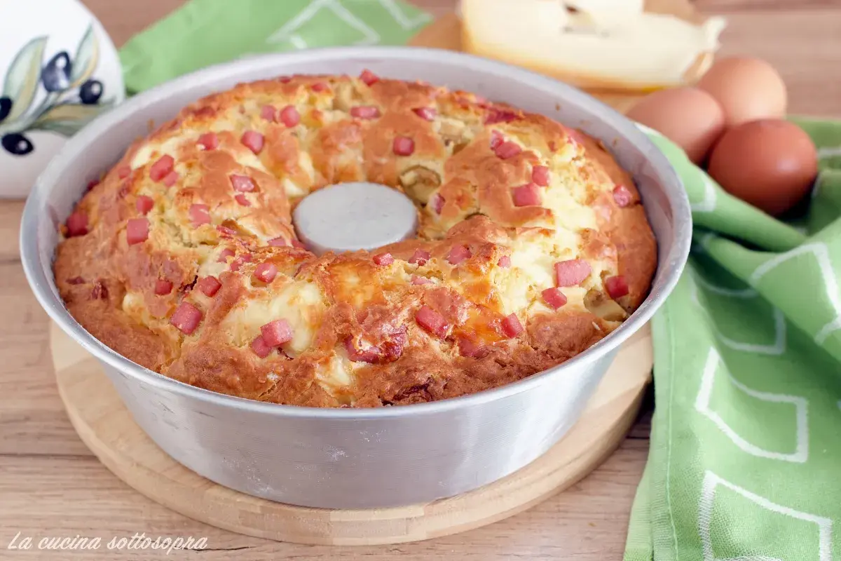 Gâteau salé végétarien doré, parsemé de dés de jambon et de fromage, dans un moule à savarin. Des œufs et du fromage sont visibles en arrière-plan.