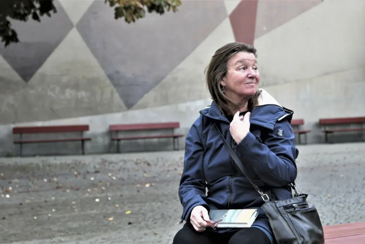 Elisabeth Rynell sitter på en bänk med en bok i knät, blickar ut mot en geometrisk vägg.