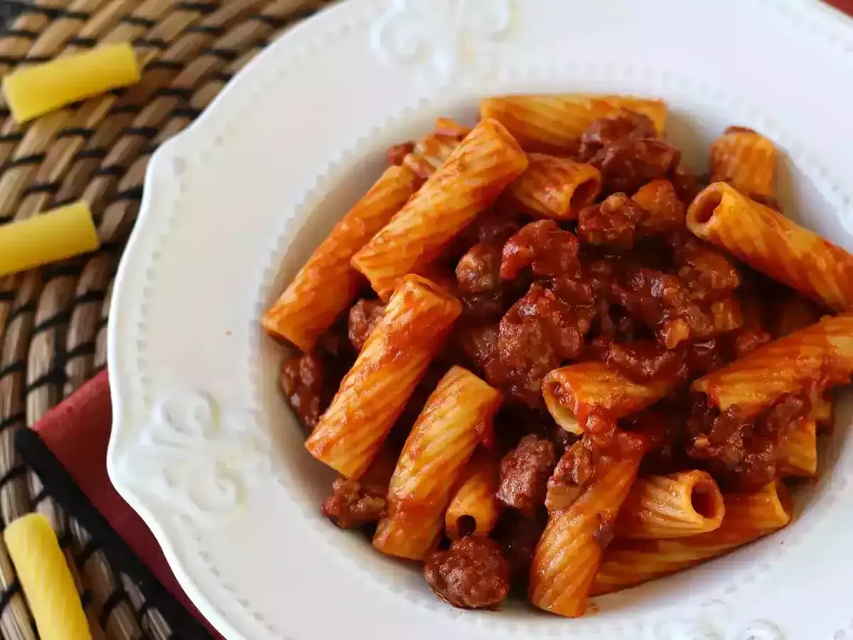 Piatto di pasta con salsiccia e sugo rosso, servita in un piatto bianco decorato.