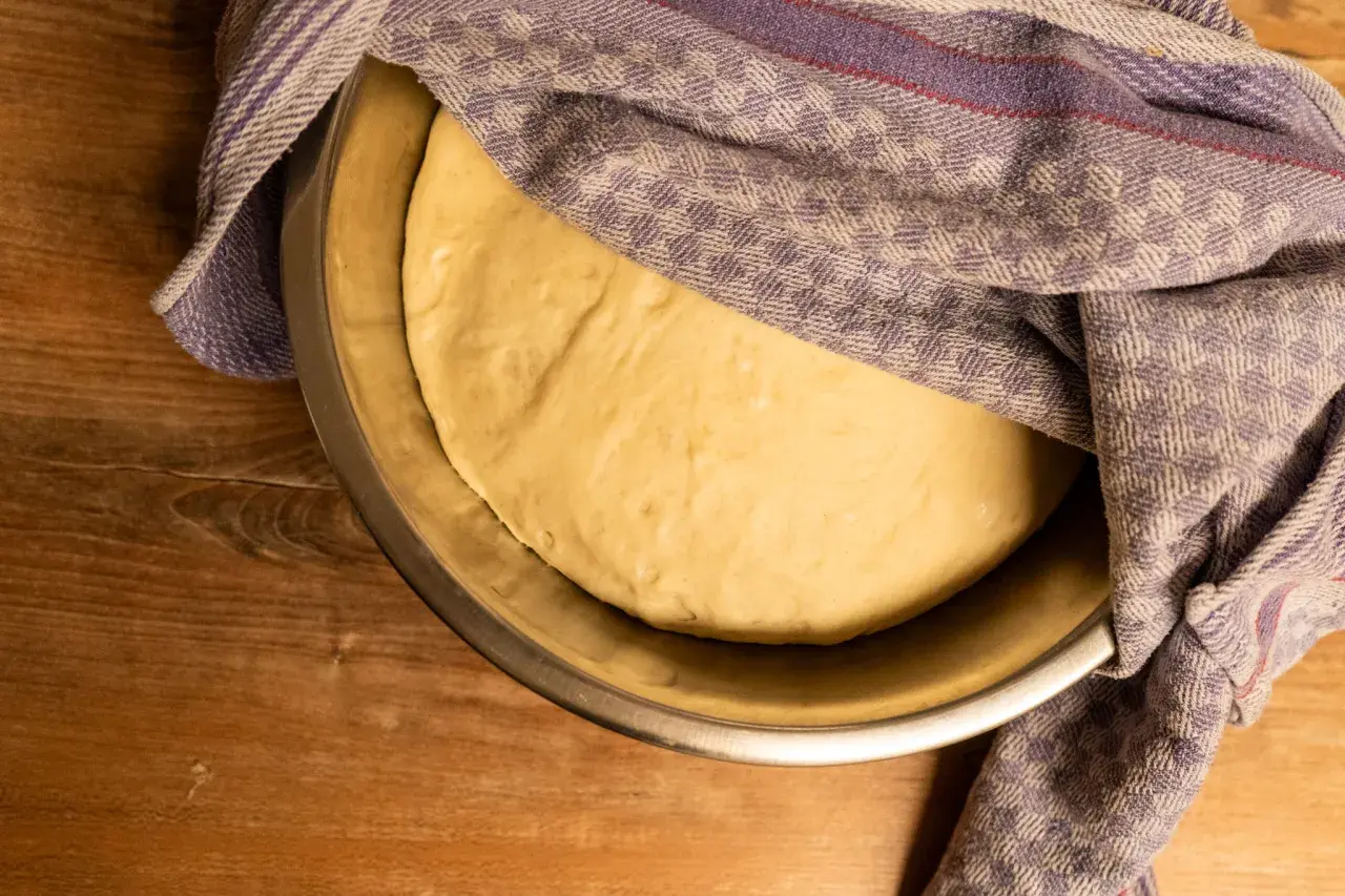 Ein goldgelber Pizzateig ruht in einer Schüssel, bedeckt mit einem karierten Tuch. Seine weiche, elastische pizzateig konsistenz verspricht eine köstliche Pizza.
