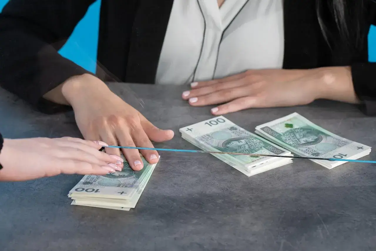 Dwie osoby przy blacie, jedna przekazuje drugiej stos banknotów 100 zł. Czy bank może żądać dowodu osobistego przy wpłacie?