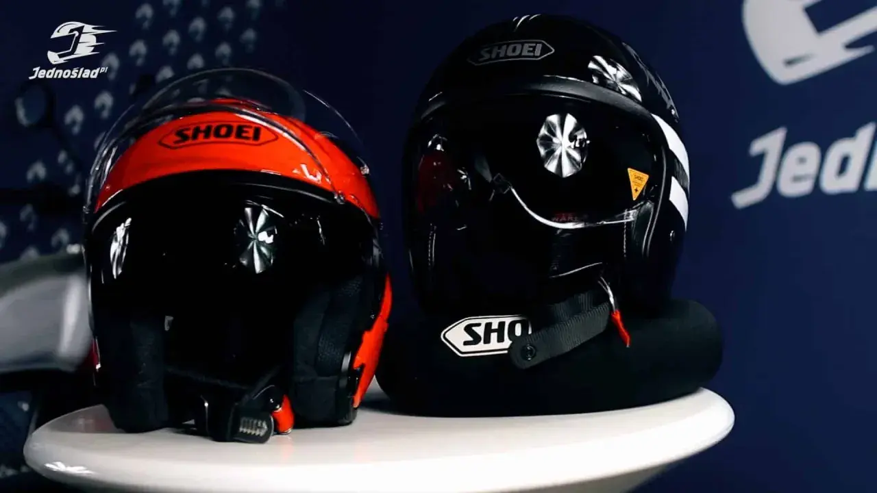 Dwa kaski motocyklowe Shoei: jeden czerwono-czarny, drugi czarny. Oba mają widoczne zapięcie kasku DD.