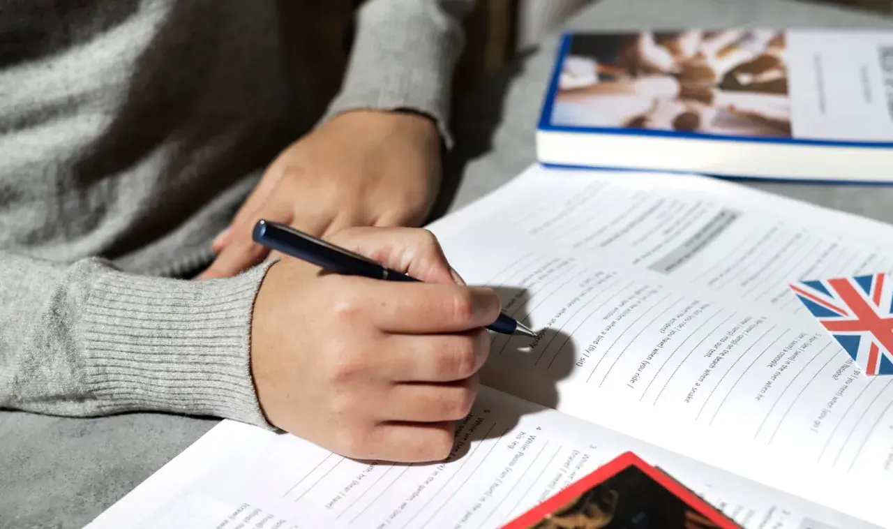 Persona escribiendo en un libro de ejercicios de inglés, con la bandera británica. Preparación para la habilitación lingüística Madrid.