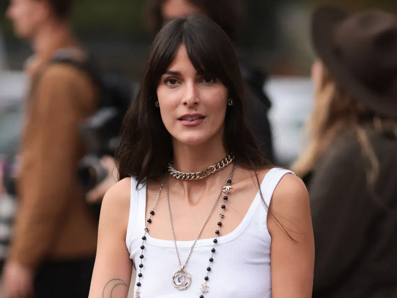 Frau mit dunklem Haar und Curtain Bangs, die eine weiße Tanktop und Chanel-Schmuck trägt.