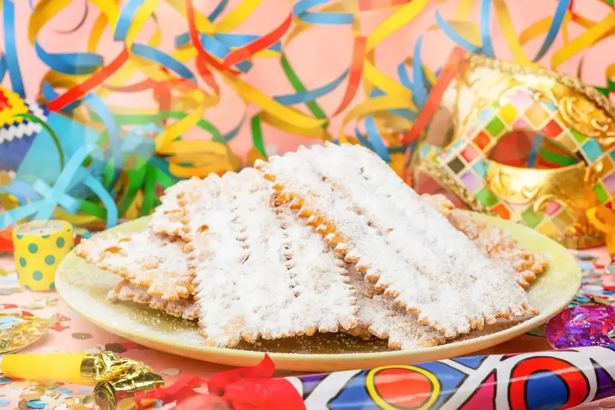 Bugie ricetta originale, dolci fritti cosparsi di zucchero a velo, su un piatto con coriandoli e maschere di Carnevale.