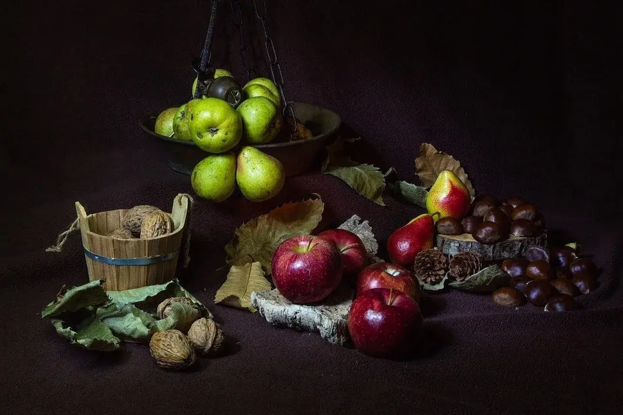 Scatti still life: mele rosse, pere verdi e rosse, noci in un cesto di legno e castagne su un tronco.