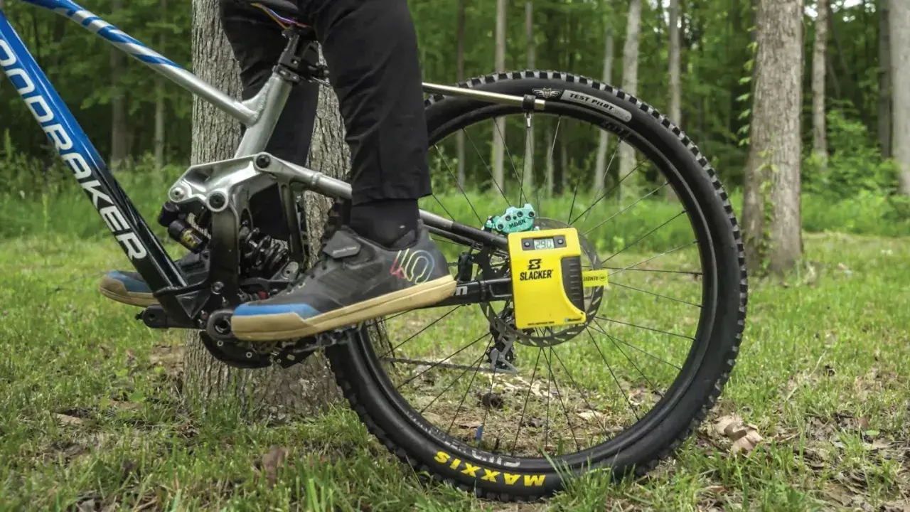 A person uses a yellow Slacker digital scale for rear shock measurements on a mountain bike, with a forest background.