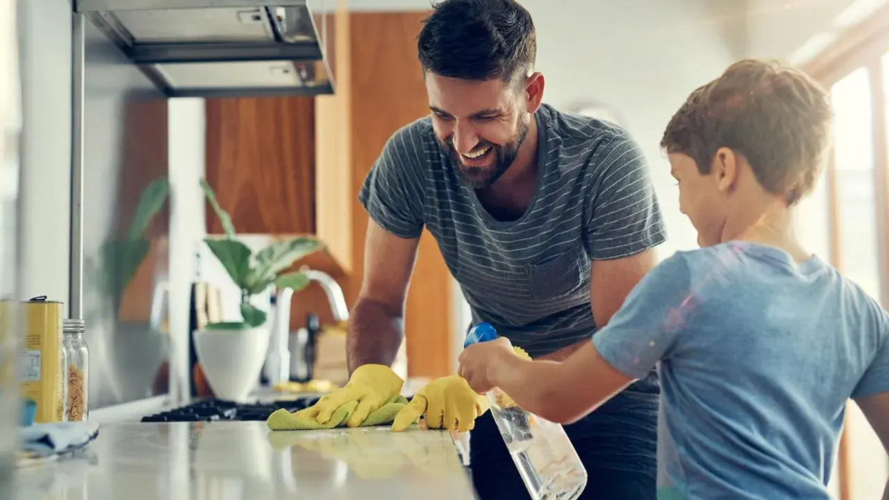 Padre e hijo ríen mientras aprenden como limpiar tu casa. El niño rocía la encimera mientras el padre la limpia con un paño.
