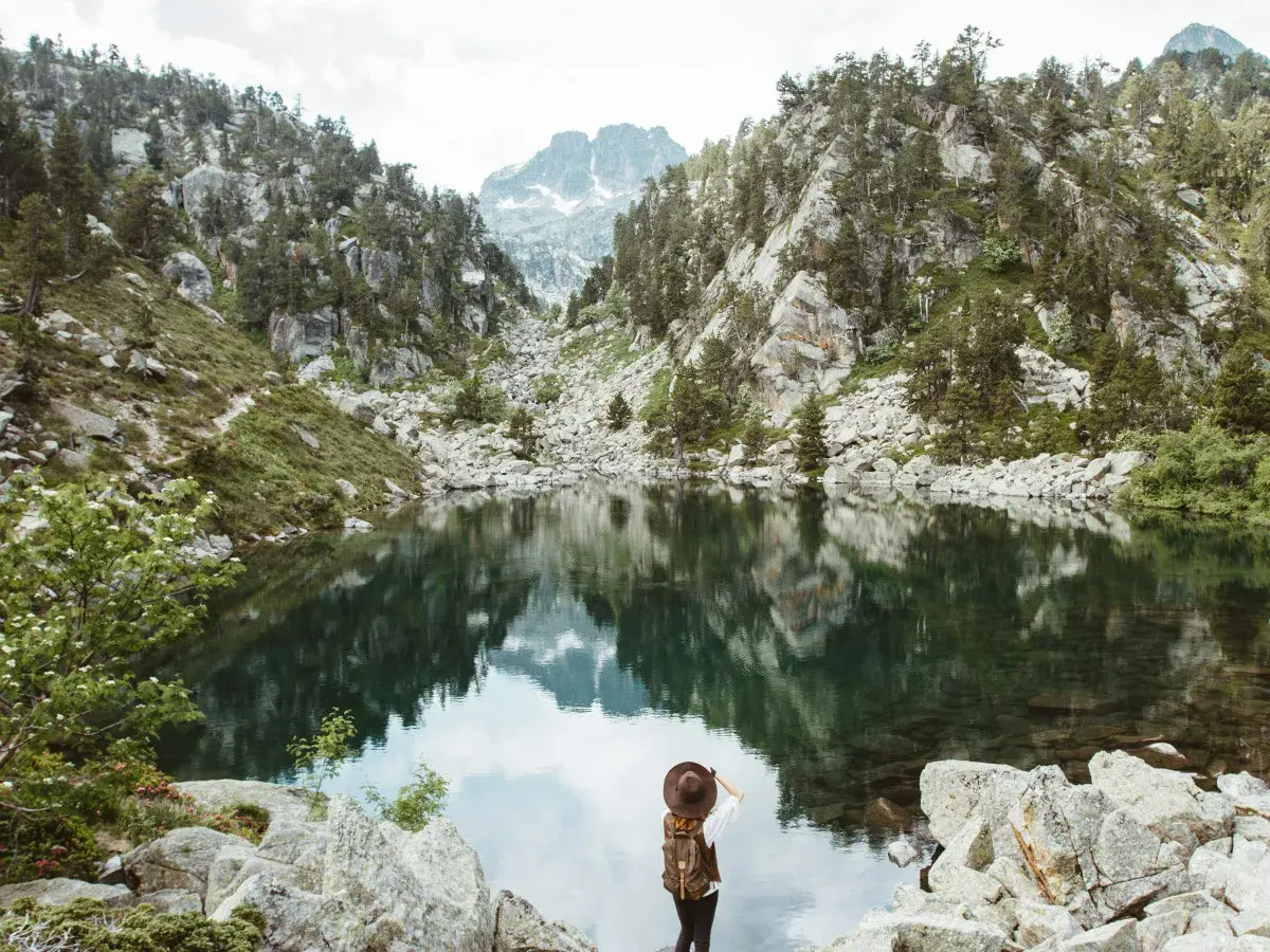 Una excursionista admira un lago sereno en el **parque nacional de los Pirineos**, rodeado de montañas rocosas y pinos.