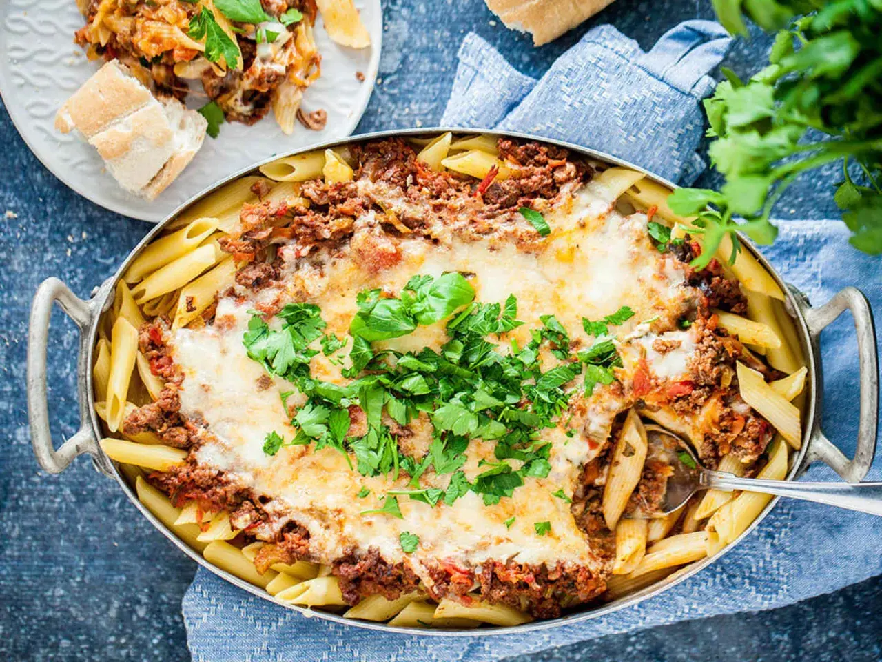 En stor ugnsform med gyllene penne, täckt med köttfärssås och smält ost. En portion pasta al forno är upplagd på en tallrik bredvid.
