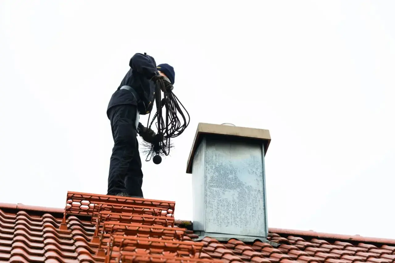 Kominiarz na dachu przygotowuje się do przeglądu komina, trzymając szczotkę i liny.
