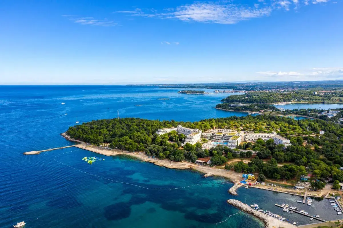 Hotel Delfin Plava Laguna z lotu ptaka, otoczony zielenią i błękitnym morzem. Widoczne baseny, plaża i przystań z łodziami.