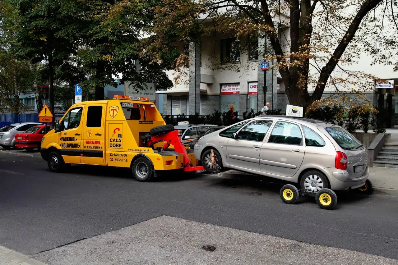 Żółty holownik odholowuje srebrny samochód. Kierowca dostał mandat za parkowanie.