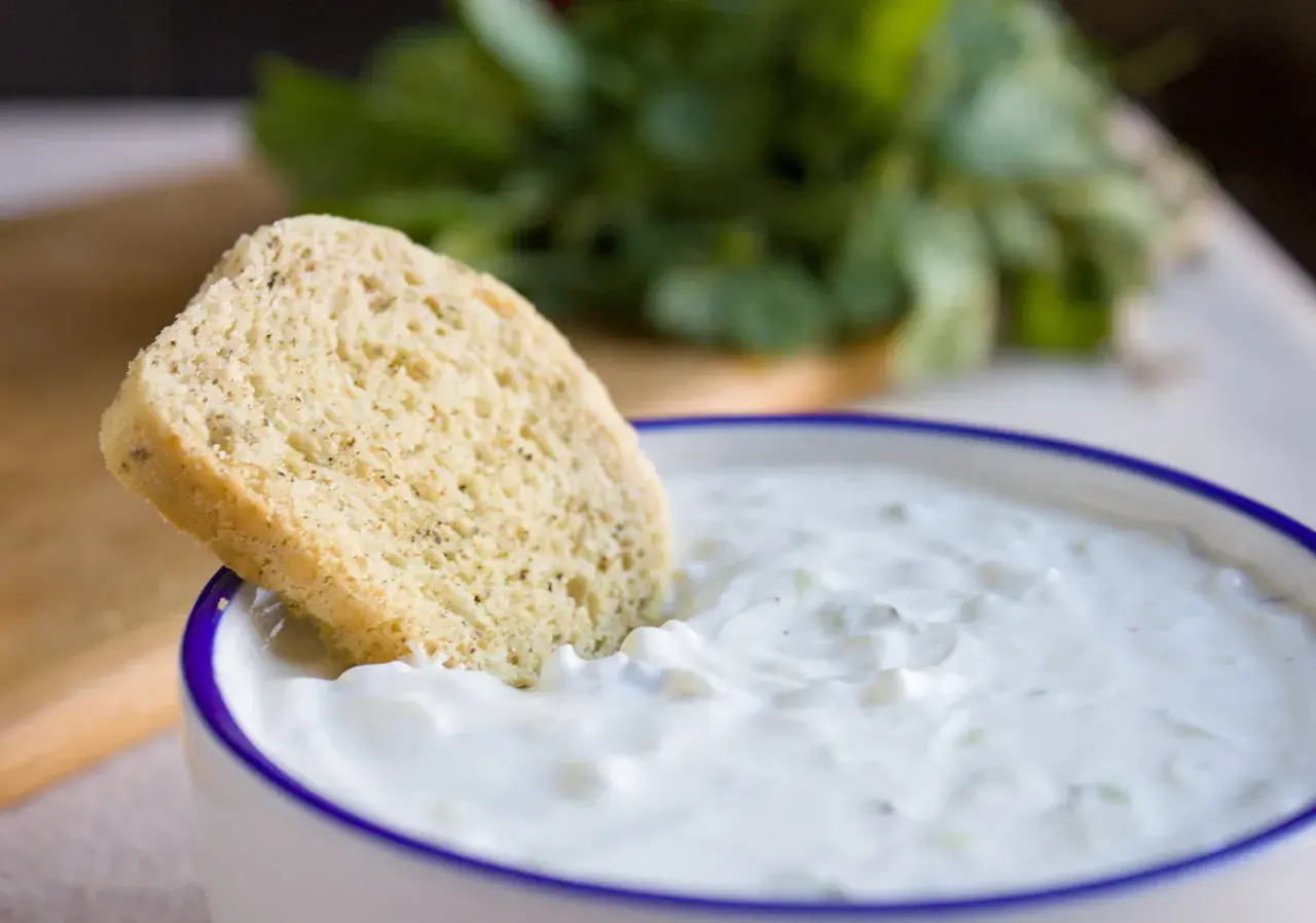 Ein Stück Brot wird in eine Schüssel mit cremigem, selbstgemachtem Zaziki getunkt. Frische Kräuter im Hintergrund.